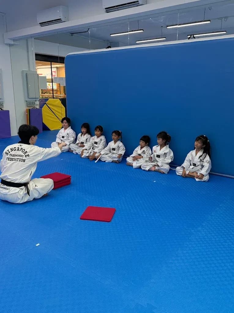 Young students in dobok sitting cross-legged in a row on the mats, listening as their instructor demonstrates.